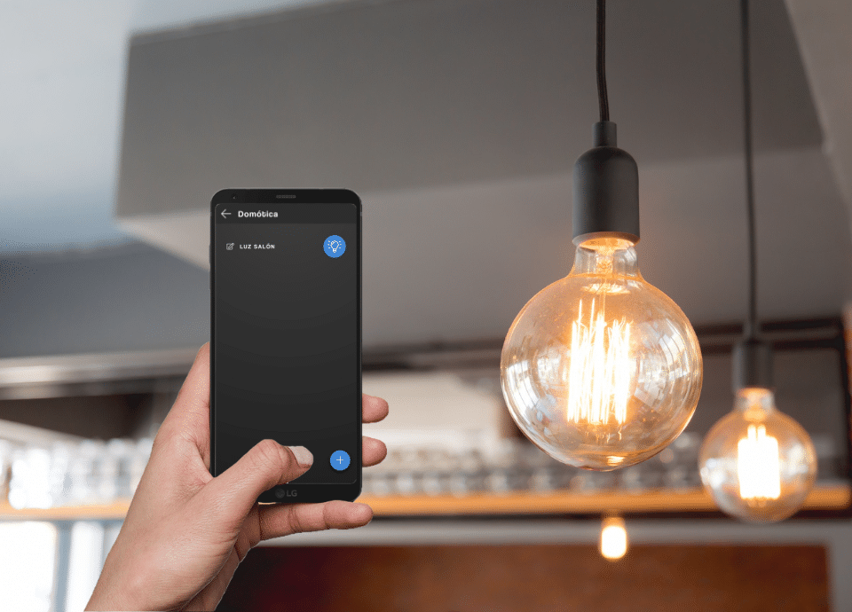 Móvil mostrando la pantalla de la app dómotica de Qvadis y encendiendo la luz de una bombilla a su derecha