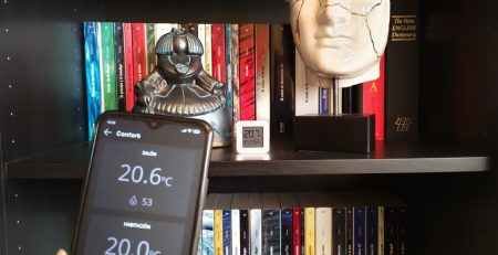 Móvil en primer plano con la pantalla de temperaturas de la app Qvadis y al fondo un termómetro Xiaomi Mijia en una estantería de un salón