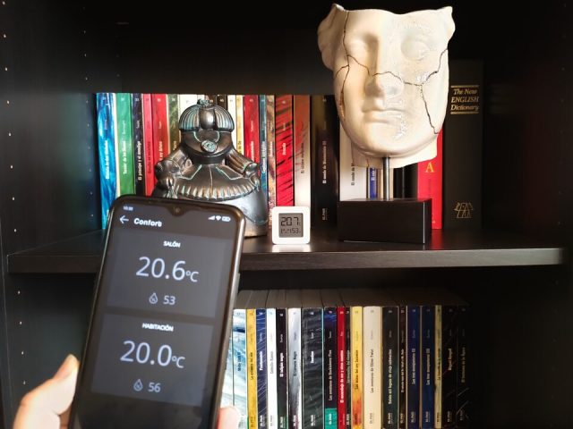 Móvil en primer plano con la pantalla de temperaturas de la app Qvadis y al fondo un termómetro Xiaomi Mijia en una estantería de un salón